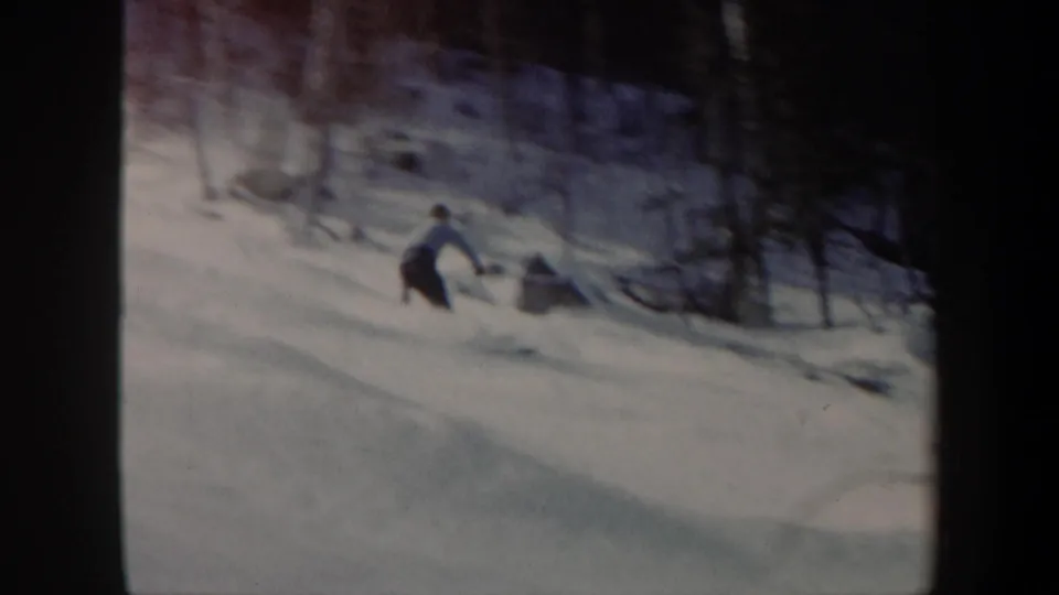 The skier continues downhill toward the right side of the frame, with bare trees and uneven snow visible in the background.