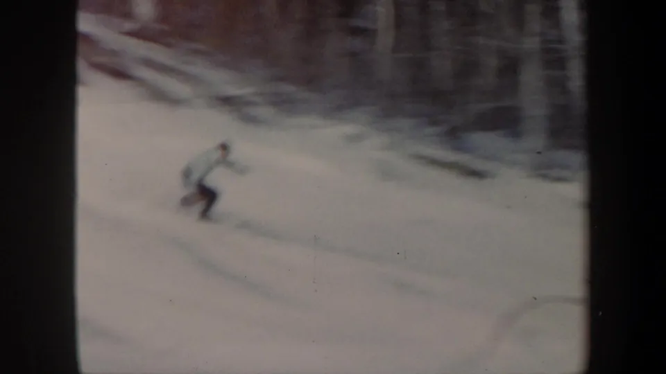 1962 Vermont home-movie frame with a blurred skier carving lower on a snowy wooded slope.
