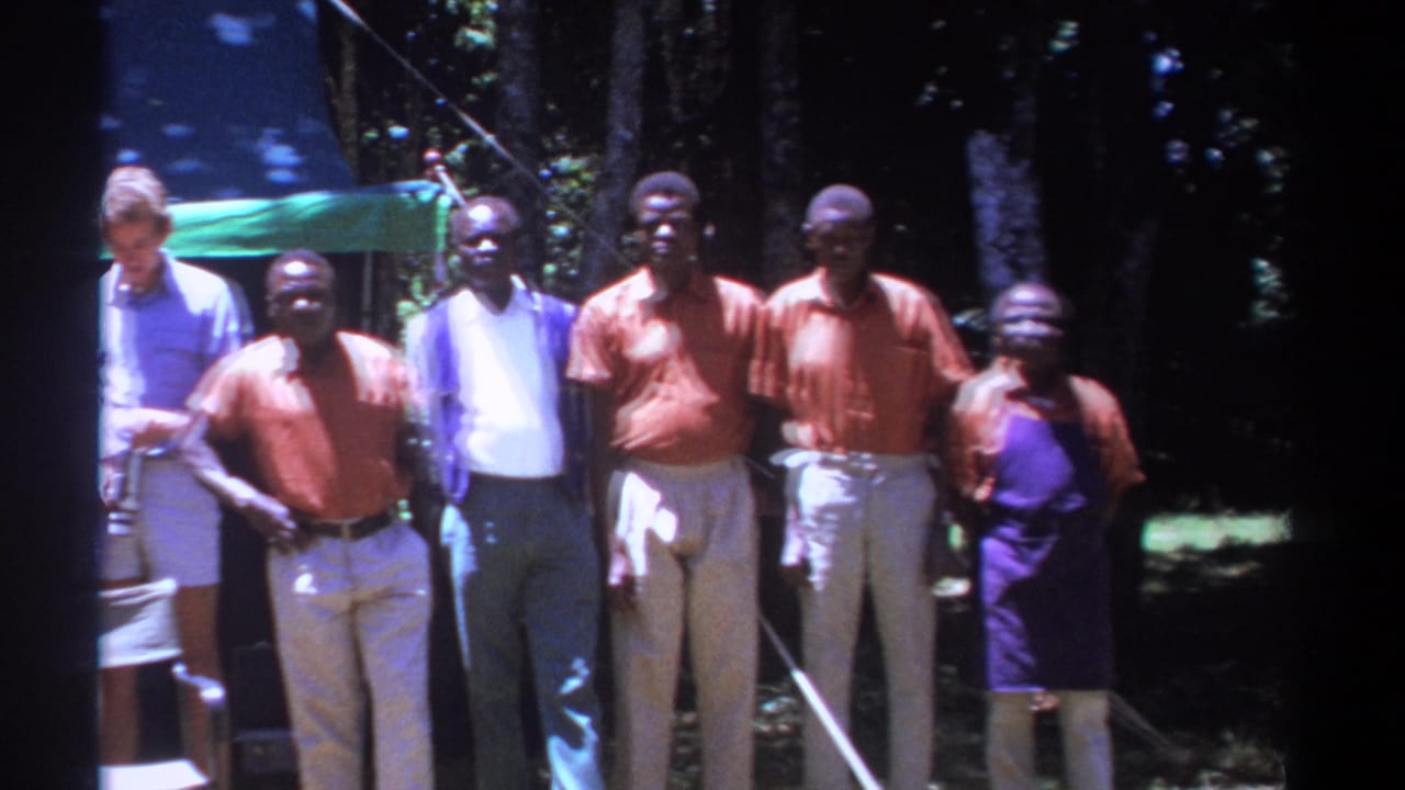 Adult men stand outdoors near a tarp shelter in a wooded area.