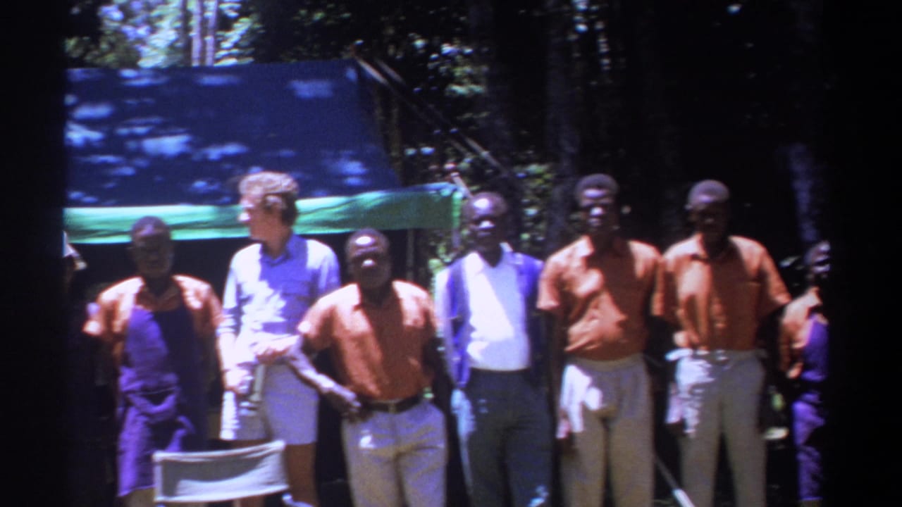 In 1969 Kenya, men in orange shirts and light trousers stand with a white visitor near a tarp shelter and folding chair.