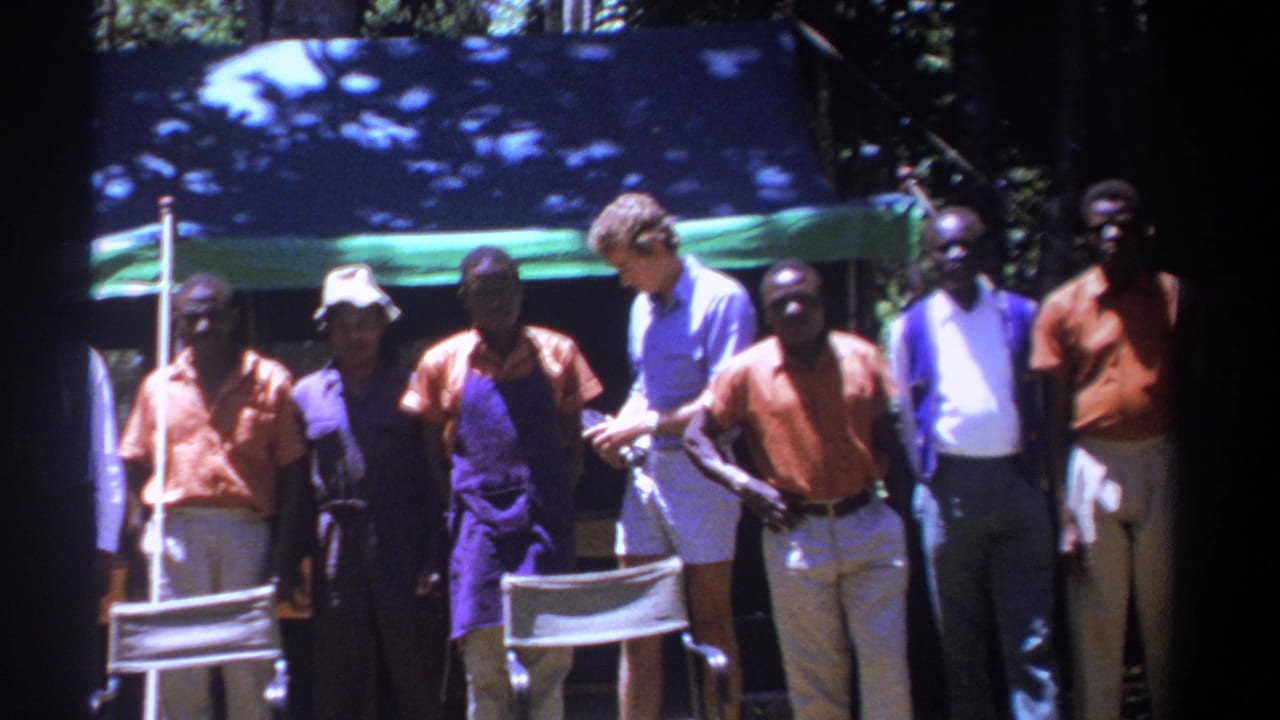 In 1969 Kenya, adults stand by folding chairs as a white man in shorts looks down at an item he is holding.