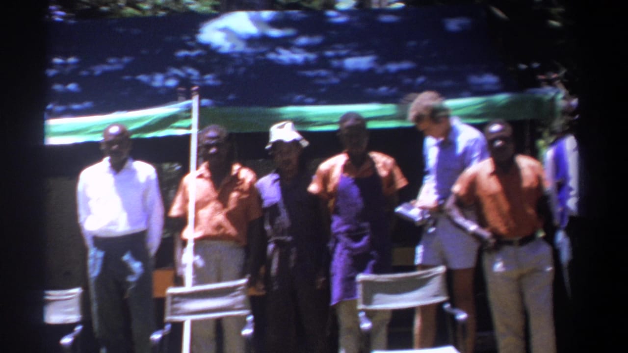 In 1969 Kenya, adult men stand in shaded daylight near a tarp shelter, with folding chairs in the foreground.