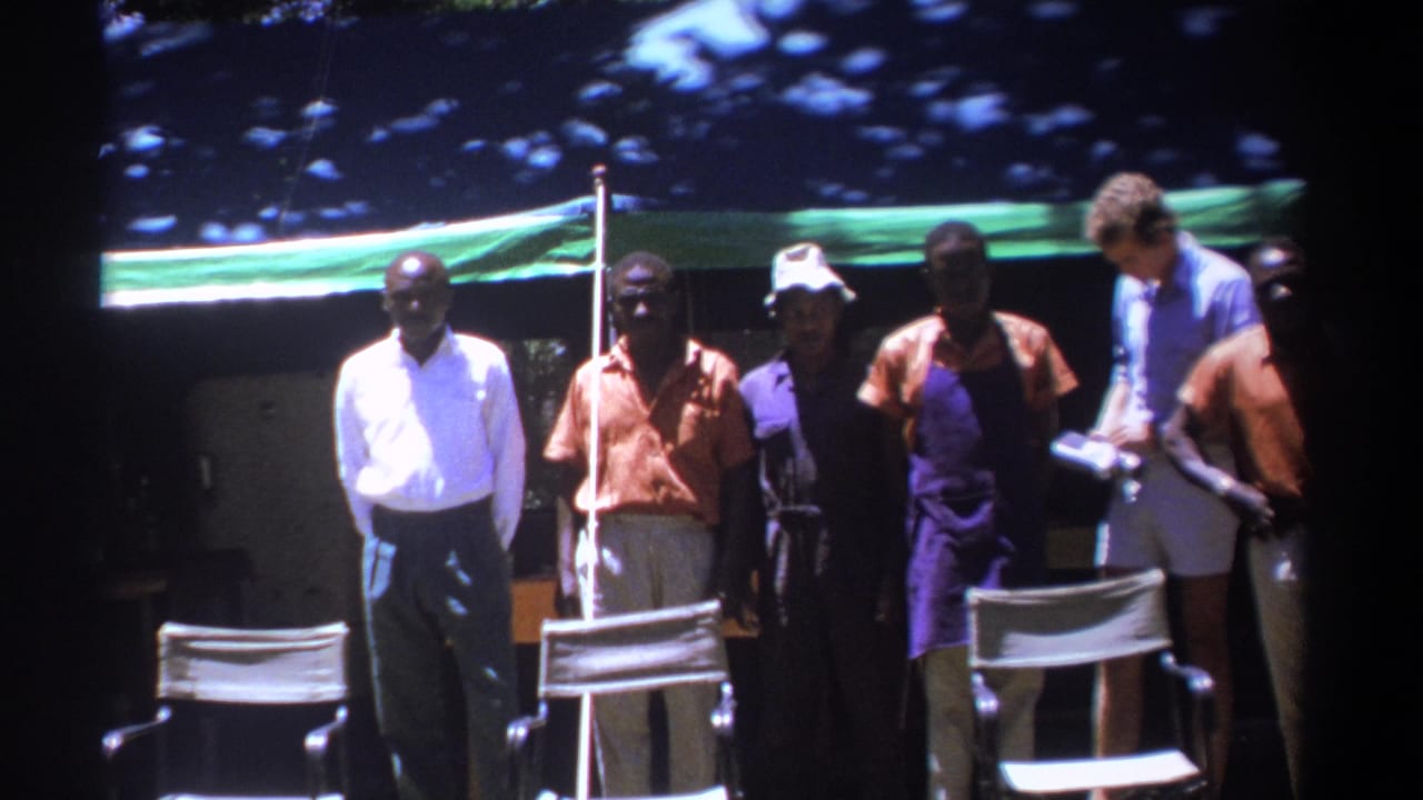 Several adult men in 1960s clothing stand in shade under a green tarp in Kenya, with folding chairs in front.