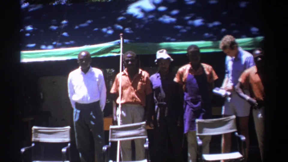 Several adult men in 1960s clothing stand in shade under a green tarp in Kenya, with folding chairs in front.