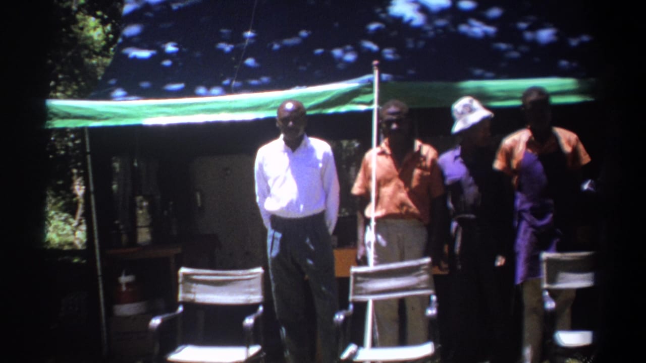 Men stand under a tarp while a light-shirted man looks down near equipment.