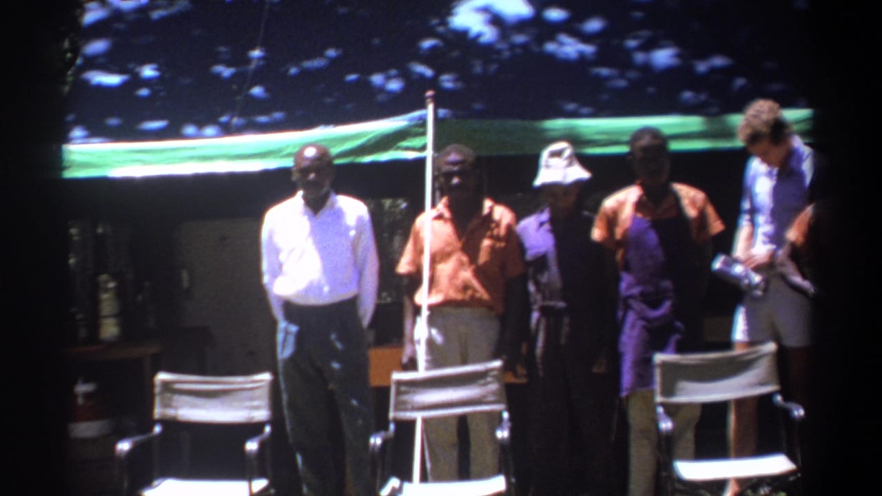 Men in shirts and trousers stand beneath a green tarp in Kenya, with director-style chairs visible in the foreground.