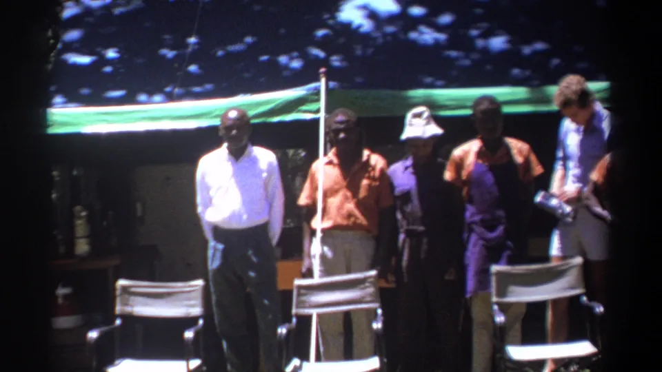 Men in shirts and trousers stand beneath a green tarp in Kenya, with director-style chairs visible in the foreground.