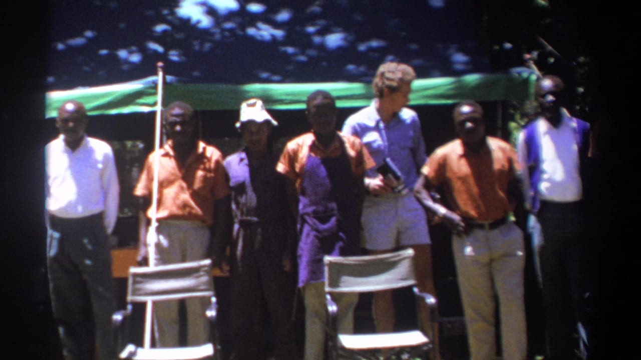 Adult men under a green tarp in Kenya face the camera while a light-shirted man stands among them with equipment or papers.