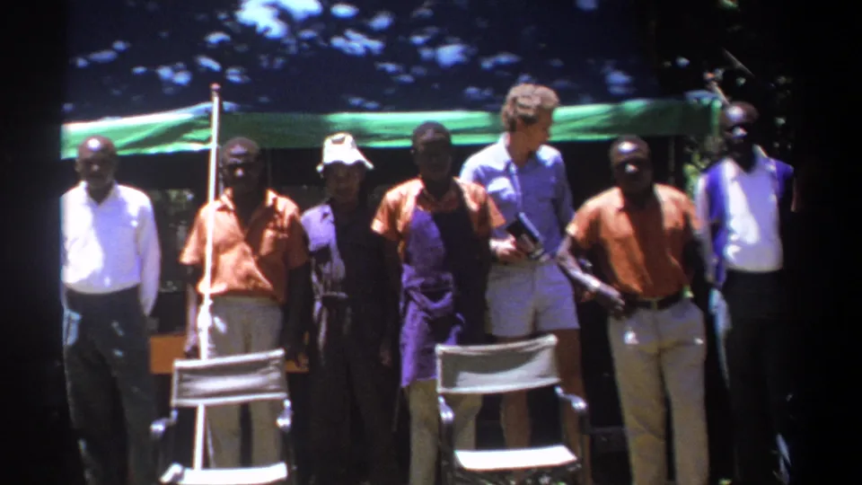 Adult men under a green tarp in Kenya face the camera while a light-shirted man stands among them with equipment or papers.