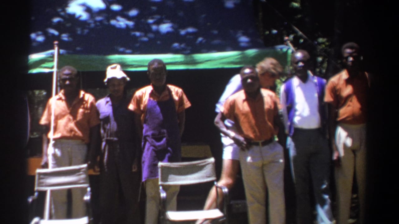 In Kenya in 1969, a group of adult men stands under trees near canvas chairs and a tarp, with several wearing matching orange shirts.