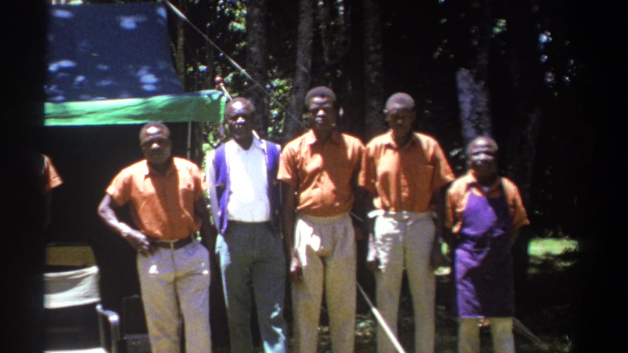 In Kenya in 1969, five adult men stand in dappled sunlight near a canvas chair, trees, and a green-and-blue tarp.