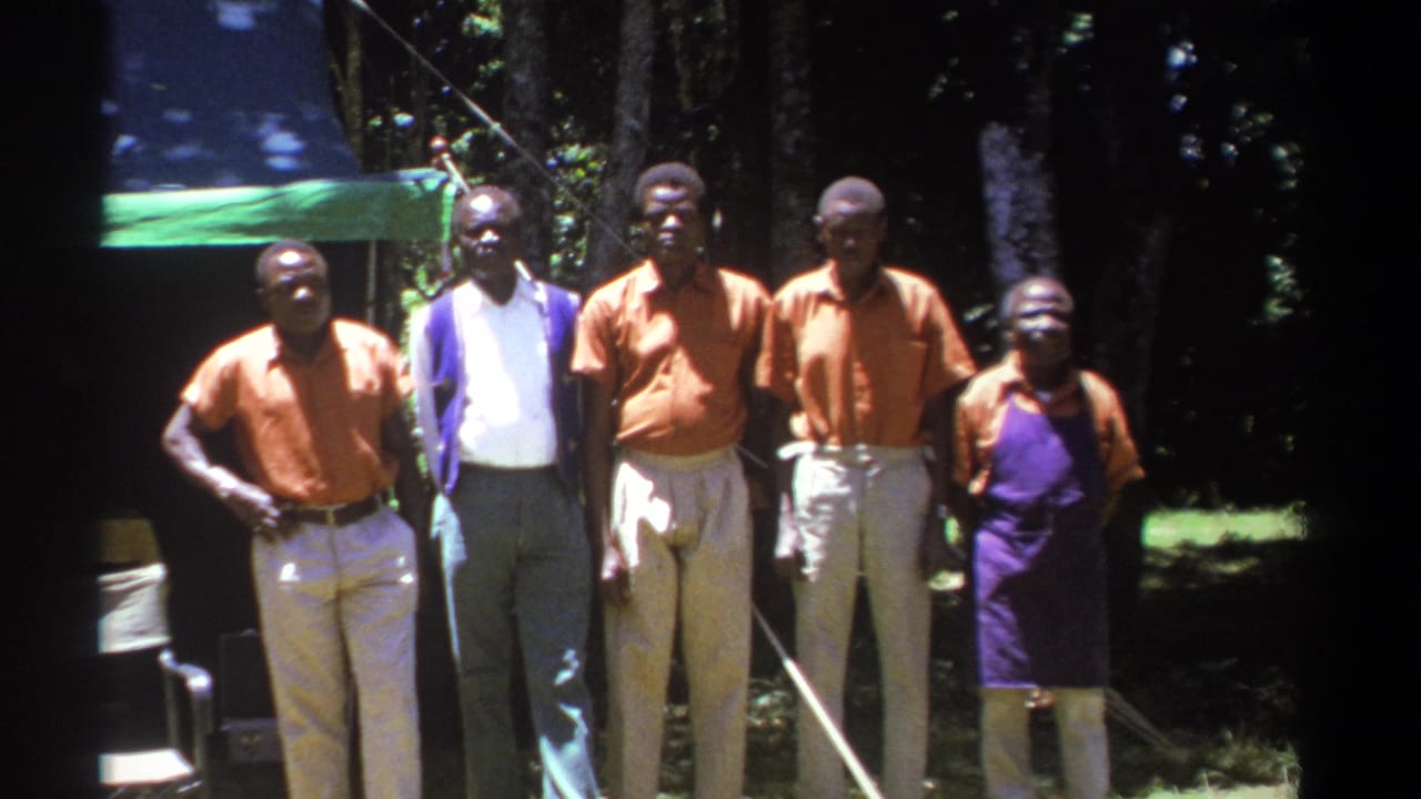 In Kenya in 1969, men in orange shirts and light trousers stand with another man in a white shirt near a tarp and wooded backdrop.