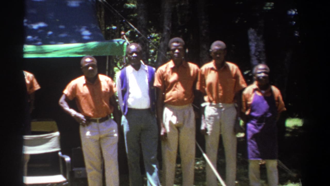 1969 Kenya 8mm frame showing adult men posing outdoors before a green tent, with trees and folding chairs visible.