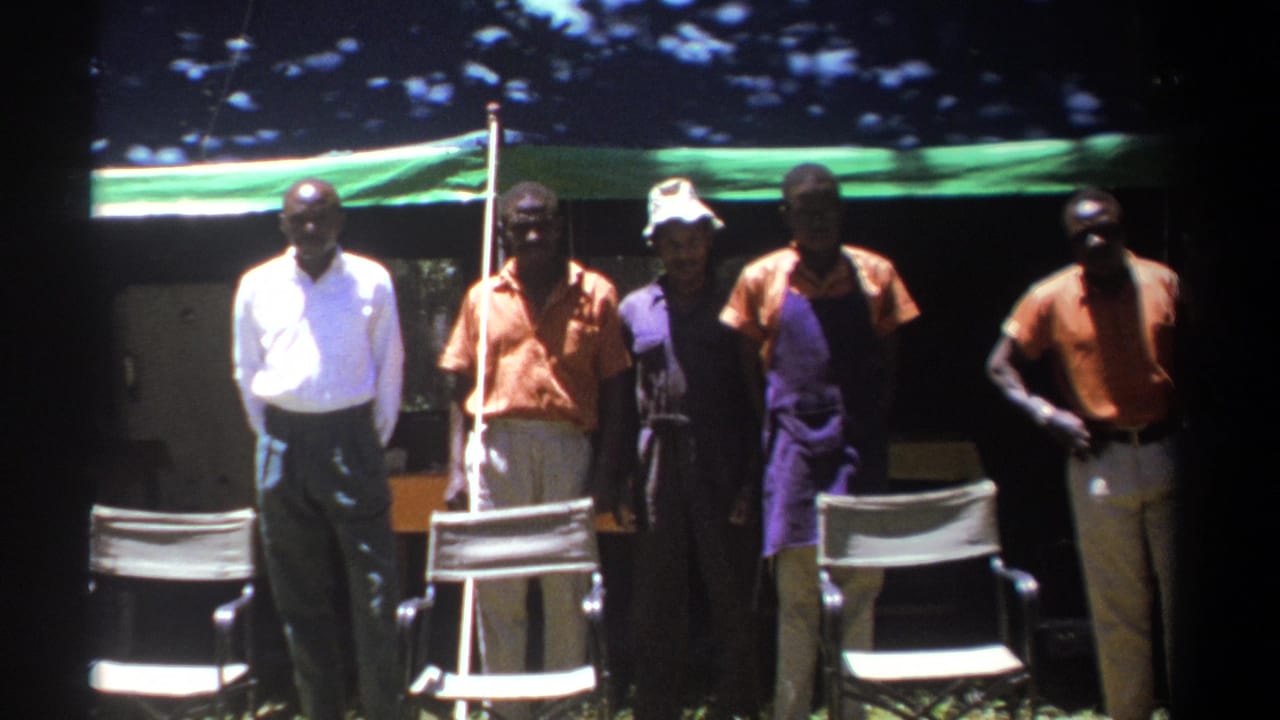 1969 Kenya home-movie frame of adult men standing by a green tent, with a white chair and wooded background visible.