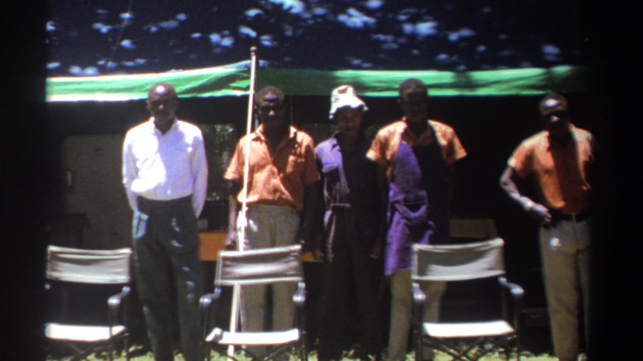 In 1969 Kenya home-movie footage, five adult men face the camera outdoors beside folding chairs and a green tarp shelter.