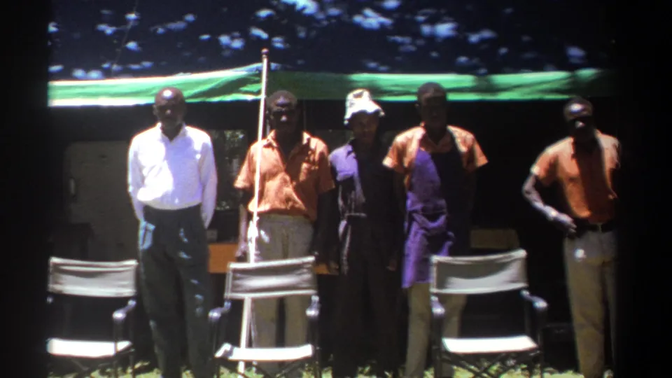 In 1969 Kenya home-movie footage, five adult men face the camera outdoors beside folding chairs and a green tarp shelter.