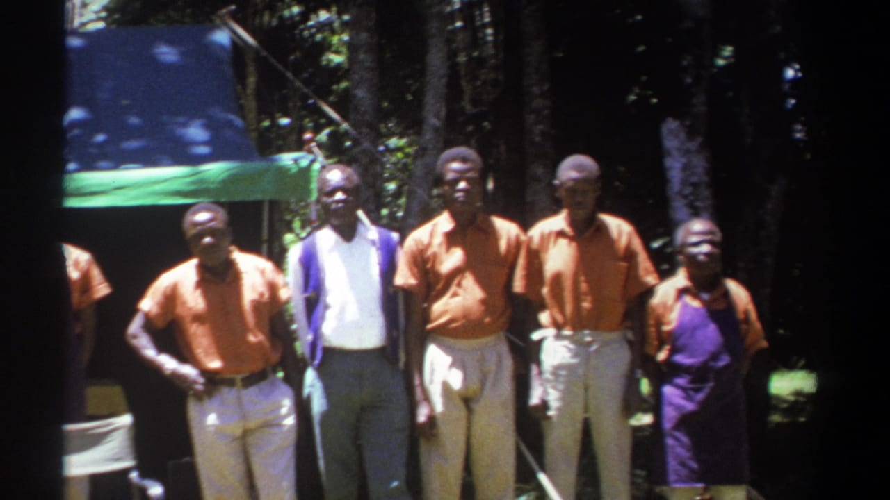 A line of adult men in 1969 Kenya stands outdoors near trees and a green tarp, with dark 8mm frame edges visible.