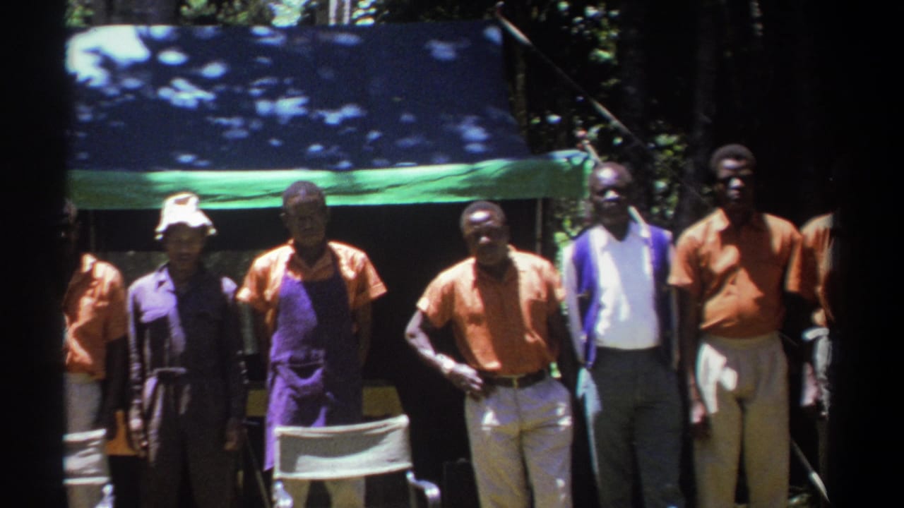 Several adult men in 1969 Kenya face the camera outdoors, with a folding chair, tarp, trees, and strong sunlight visible.