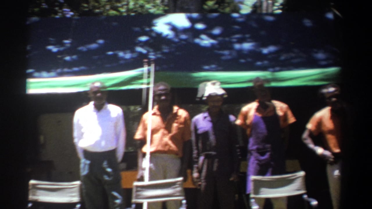 Adult men in 1969 Kenya stand under or near a green tarp, with folding chairs in front and wooded shade behind them.