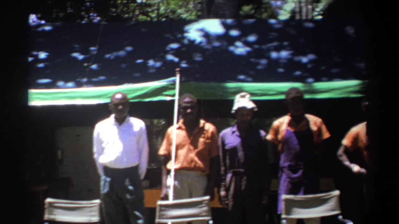 1969 Kenya 8mm frame of several men standing beneath a green-and-blue tarp among trees, with chairs partly visible in front.