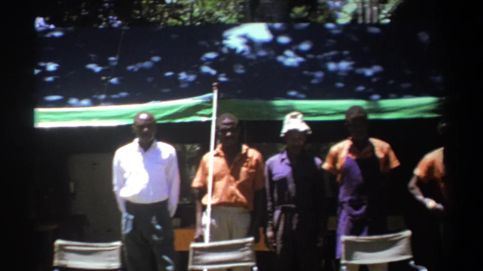 1969 Kenya 8mm frame of several men standing beneath a green-and-blue tarp among trees, with chairs partly visible in front.