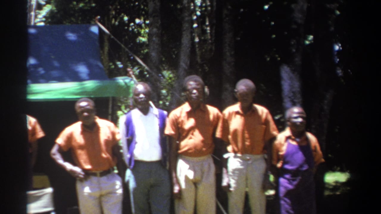 1969 Kenya 8mm frame of a small group of men in orange shirts and aprons standing near folding chairs in a forested setting.