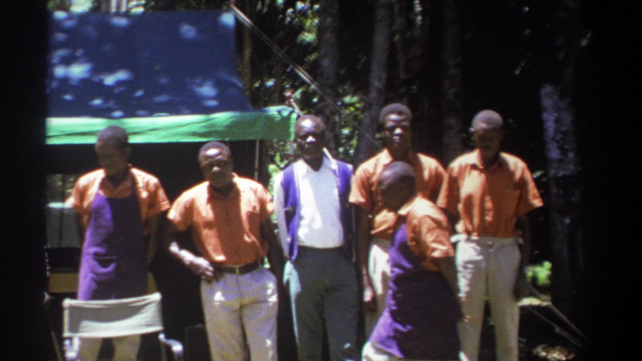 In Kenya 1969 home-movie footage, a group of adult men in matching work clothing stands beneath a tarp among trees.