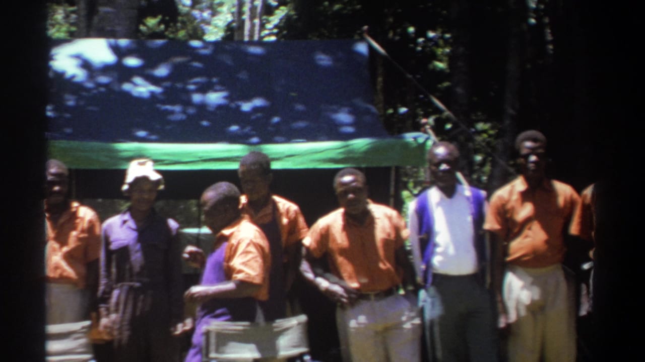Adult men in Kenya stand near a shaded tarp and trees, some in orange shirts and one in a white hat, in 1969 8mm footage.