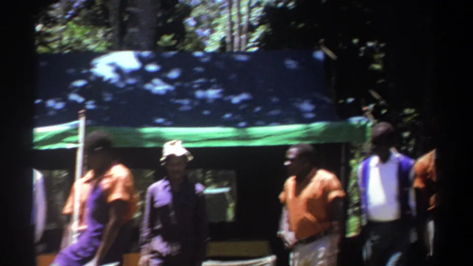 In the final frame, one man moves across the foreground while other adult men remain gathered under the tarp in wooded Kenya footage.