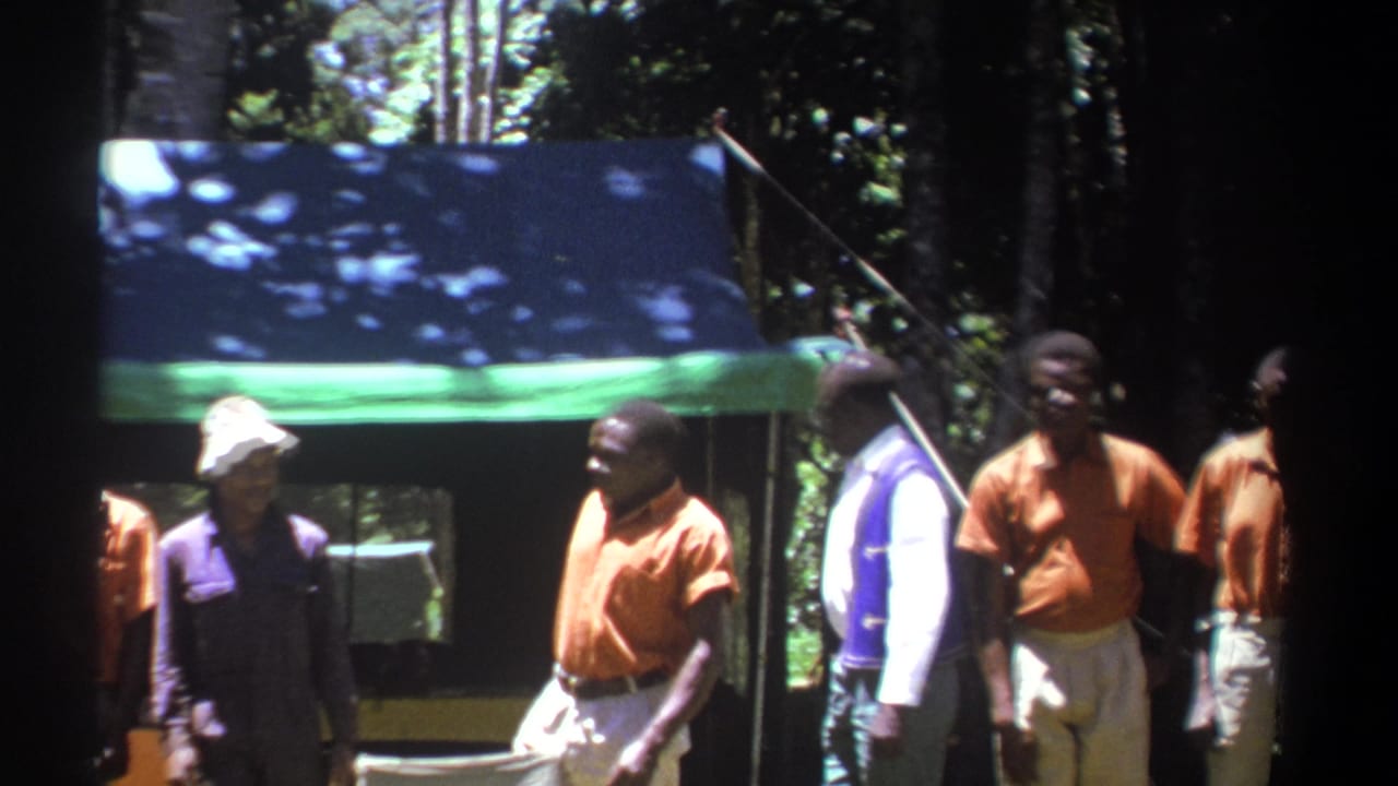 In 1969 Kenya footage, several men gather beside a blue-and-green tent under trees, with bright orange shirts visible.