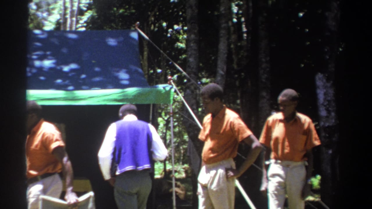 Men in orange shirts and one man in a purple vest stand beside a tent.