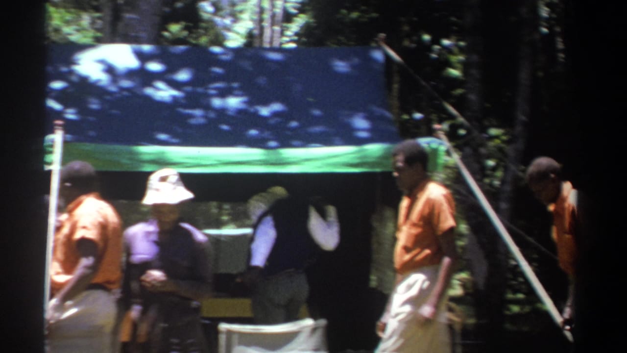 In 1969 Kenya footage, people walk and stand around chairs beside a blue-and-green tent in a wooded clearing.