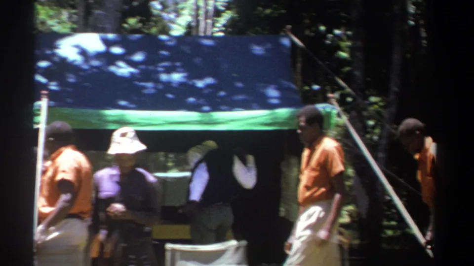 In 1969 Kenya footage, people walk and stand around chairs beside a blue-and-green tent in a wooded clearing.