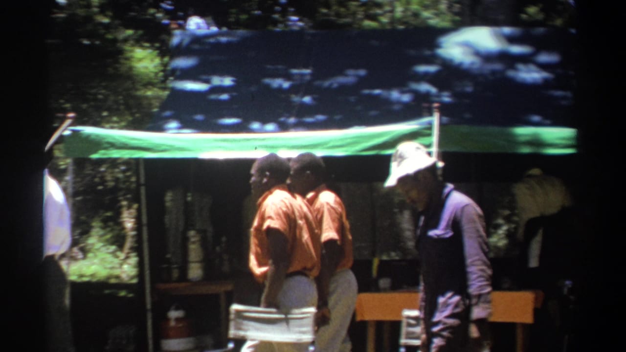 In 1969 Kenya footage, men in orange shirts and a man in a purple vest stand near a tent with chairs and items beneath it.