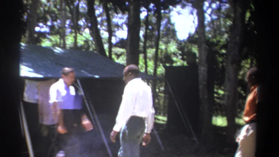 1969 Kenya 8mm frame with men in white shirts walking beside a large canvas tent in a wooded campsite.