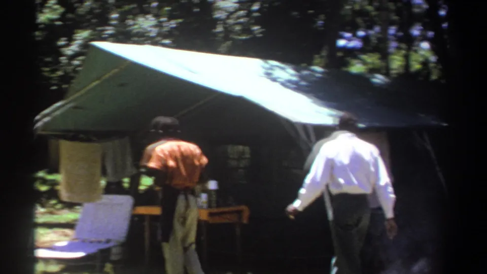 1969 Kenya 8mm frame showing men near a green canopy tent, folding chair, and towel in a wooded camp area.