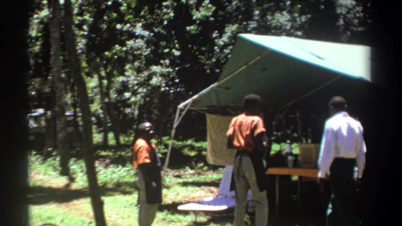 1969 Kenya 8mm frame showing two men by a green canopy tent with trees and camp items in the background.