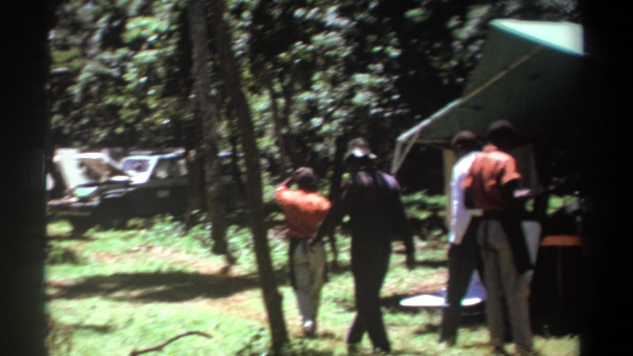 1969 Kenya 8mm frame showing men walking near a green canopy tent, with trees, grass, and a low cot or bench in the camp area.