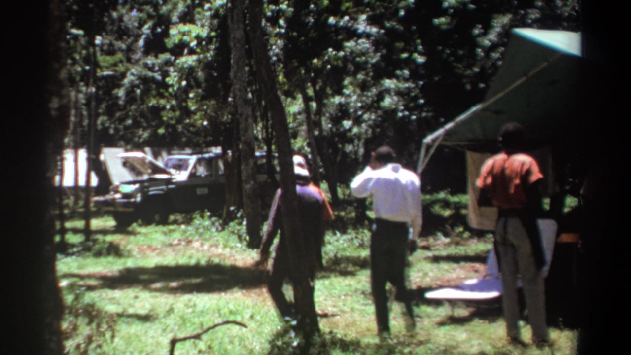 1969 Kenya 8mm frame showing men walking through a forest campsite near a canopy tent, with a dark vehicle partly visible beyond the trees.
