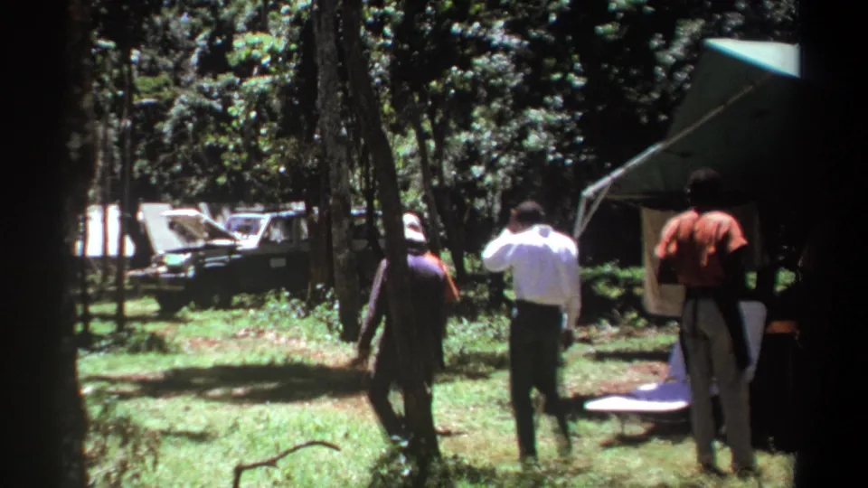 1969 Kenya 8mm frame showing men walking through a forest campsite near a canopy tent, with a dark vehicle partly visible beyond the trees.