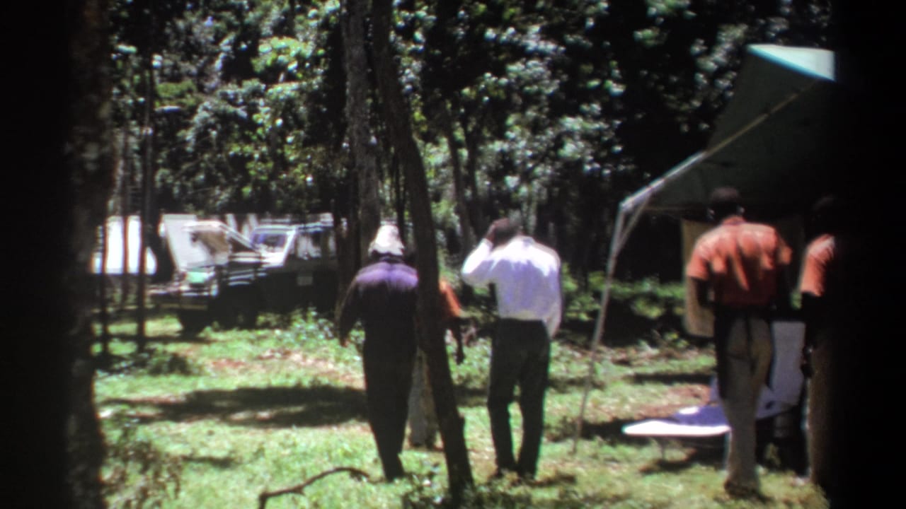 In 1969 Kenya home-movie footage, several men walk away through a wooded campground toward a canvas tent and a vehicle with its hood open.