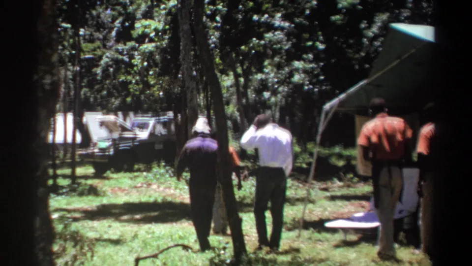 In 1969 Kenya home-movie footage, several men walk away through a wooded campground toward a canvas tent and a vehicle with its hood open.