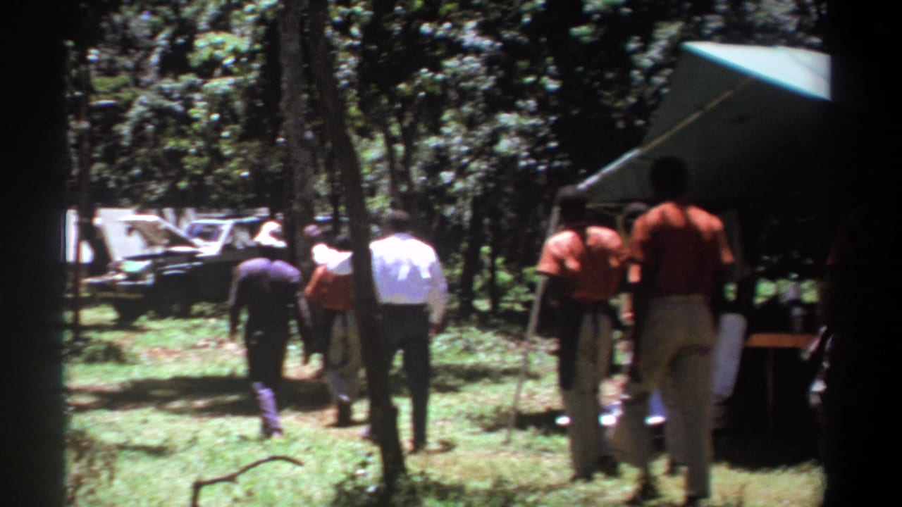 A group of men continues away from the camera beside a tent in a wooded clearing.