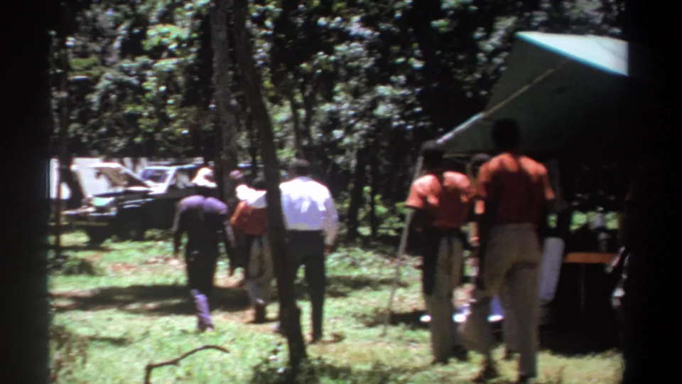 The group moves farther into the shaded campsite, with men in orange shirts and light trousers near a canvas tent and vehicle.