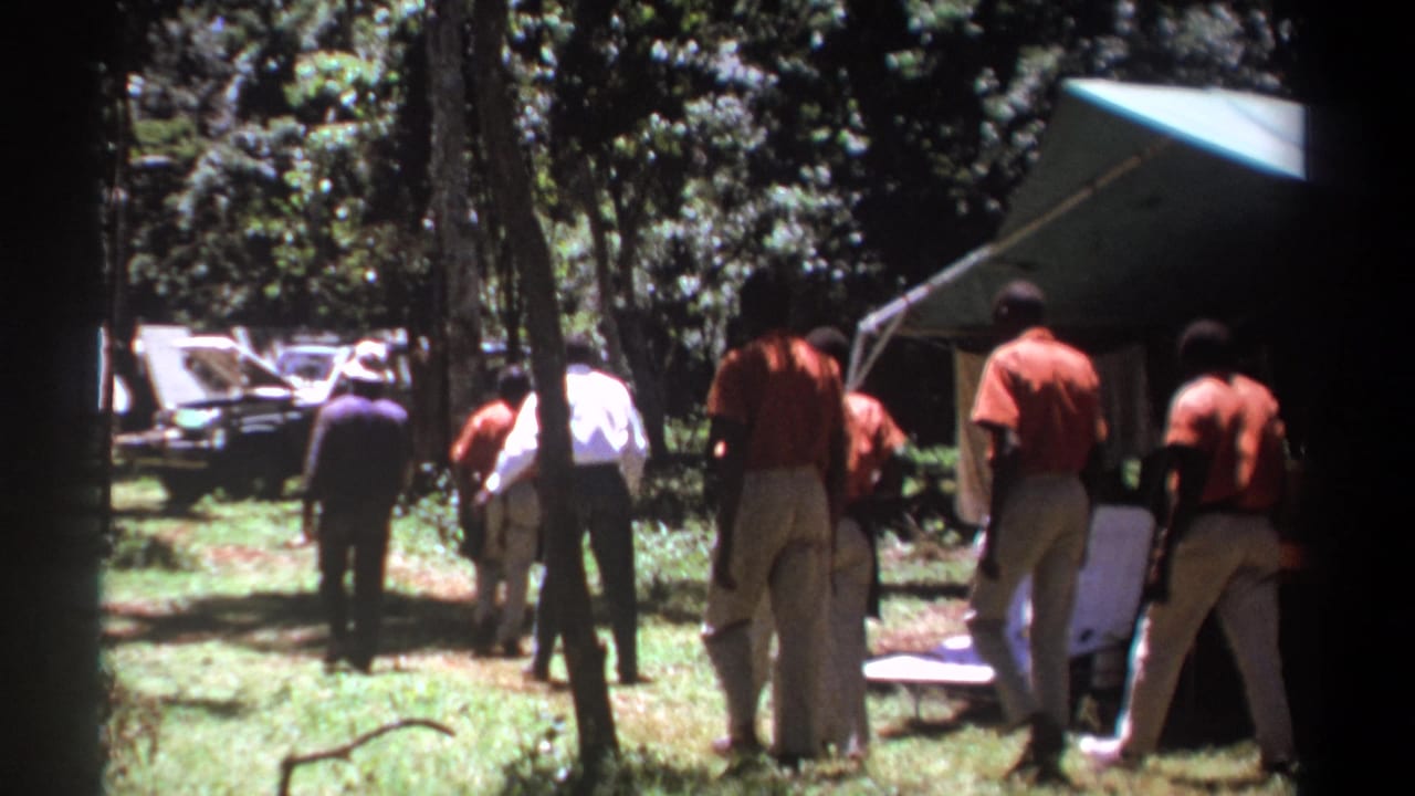 In the wooded Kenya campsite, the men continue walking away, passing near folding chairs, a tent, and a vehicle in the background.