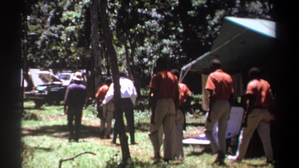 In the wooded Kenya campsite, the men continue walking away, passing near folding chairs, a tent, and a vehicle in the background.