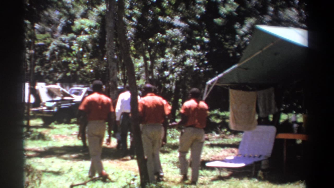 In Kenya 1969 home-movie footage, men in red and white shirts walk away through a grassy campsite beside a green canopy.