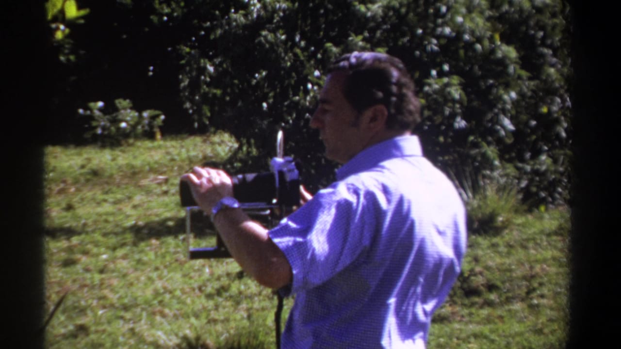 1969 Kenya home-movie frame showing an adult man in a blue shirt using a vintage camera in a sunny grassy clearing.