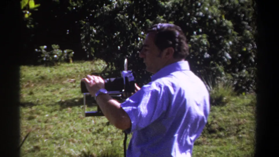 1969 Kenya home-movie frame showing an adult man in a blue shirt using a vintage camera in a sunny grassy clearing.