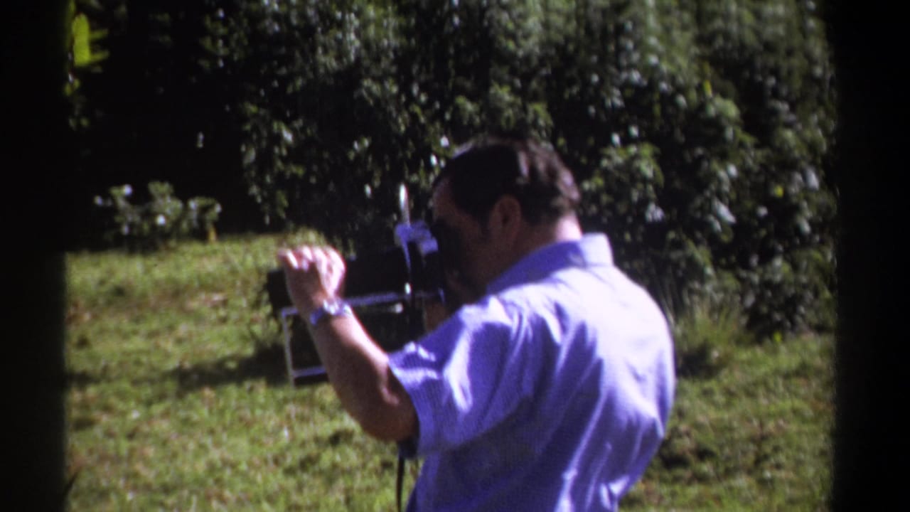 In 1969 Kenya home-movie footage, a man in a light blue shirt films outdoors with a vintage 8mm camera amid grass and trees.